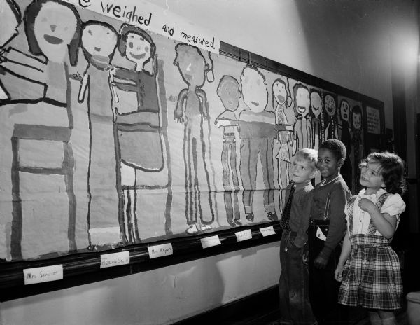 Three Franklin school second grade students looking at a mural the students made of themselves to track their weight and height. From left are: Steven Willadsen, Donald Harris, and Joannie Ranney. Their teacher is Mrs. Charlotte Severson.