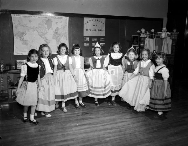 Nine Nakoma School third grade girls dressed in costumes of the countries they represented for a class play, "Democracy for Boys and Girls." The children were assigned to find out where their ancestors came from and what they brought with them to America.  Left to right: Mary Whitford, representing England; Maxine Lippoit, Germany; Diane McGovern, Ireland; Bonnie Timmons, Ireland; Bonnie Clements, Holland; Reeanne Finningan, Norway; Nancy Larson, Switzerland; June Honeck, England; and Marcia Friedman, Russia.