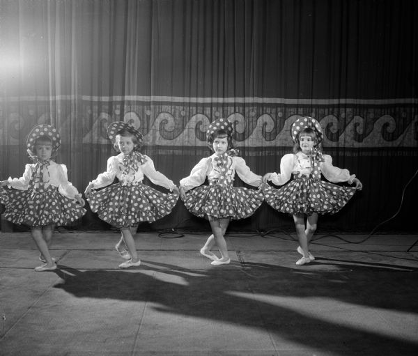 Four young girls wearing polka-dot dresses and hats are shown on stage at the Masonic auditorium. They are dancing the roles of the dolls in the 'March of the Toys' in 'The Nutcracker' ballet. They are, left to right, Janet Hartwich, Joelyn Ripp, Sussan Lee Brown, and Jacquie Harmon.