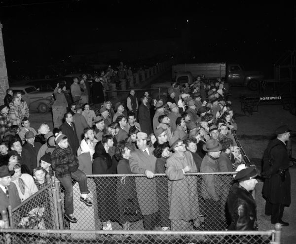 Part of a crowd of 150 waiting at the fence at the Truax airport terminal as the Badger football team's chartered airplane returns at 8:20 PM after losing to Pennsylvania 20-0. Shows the crowd, cars, trucks and a police officer with a gun in holster.