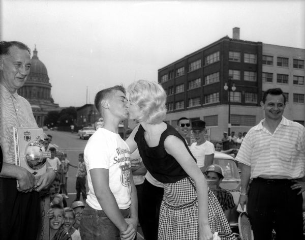 The winner of the 1957 Soap Box Derby, Van Steiner, receiving champion's kiss from Barbara Harned, wife of "Wisconsin State Journal writer, who was a former stewardess with Germany's Lufthansa Airlines. Building in the background is the Ray-O-Vac Office, 212 East Washington Avenue, and the Wisconsin State Capitol.