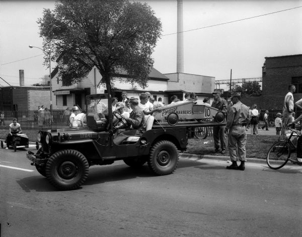 The Marine Corps Reserve built platforms on their jeeps to move the soap box cars back up the East Washington Avenue hill. They are shown loading the Portage Barbers car. They also provided field phone communications. Armour & Co. office, 633 East Washington Avenue, is in the background.