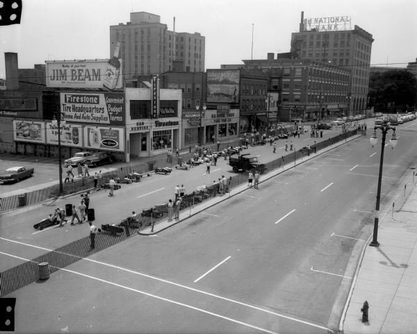 Elevated view of Soap Box Derby staging area and starting line on 100 and 200 blocks of East Washington Avenue between South Butler and South Pinckney Streets. Businesses (left to right): parking lot for First National Bank, Firestone Tires, Interstate Finance Corp., U.S. Royal Tires (Pahl Tire Co.), U.S. Army Recruiting Station, entrance to Harloff Apartments, Wagon Wheel tavern, Remington Rand business machines, (South Webster Street), Washington Army Store, entrance to the Washington Building, Kroger grocery store, Family Finance Corp, Uptown Liquor Store. A portion of the Wisconsin State Capitol is visible in the background.