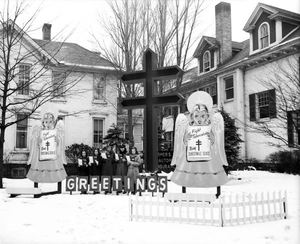 A group of girl scouts from Troop 99 of Bashford Methodist Church singing Christmas carols at a Christmas holiday display at a Madison law firm featuring the Christmas seal design of the National Tuberculosis Association. The girls in the picture, from left, are Jean Hillestad, Janet Stignani, Mary Ann Duerst, Joan Brown, and Rose Marie Thuesen.