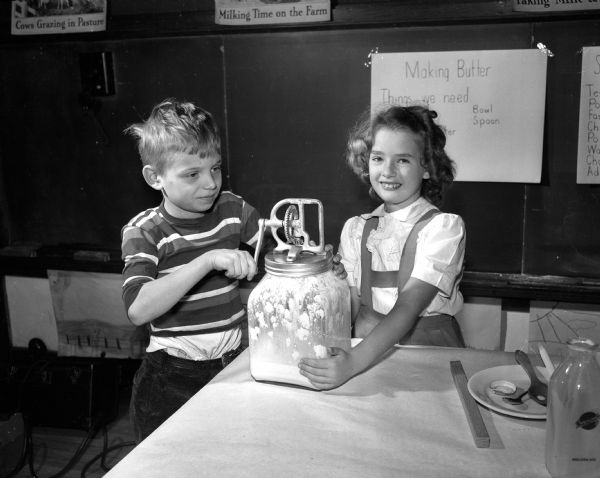 Second grade children of Lowell School engaging in a class project of making butter as part of a dairy study. Warren Oakes, (left) is churning the cream with the help of Kathleen Roman.