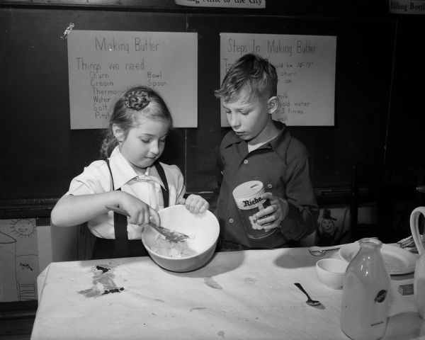 Second grade children of Lowell School engaging in a class project of making butter as part of a dairy study. Sally Myers, (left) is stirring the butter while Sammy Smith is adding a "touch" of salt.