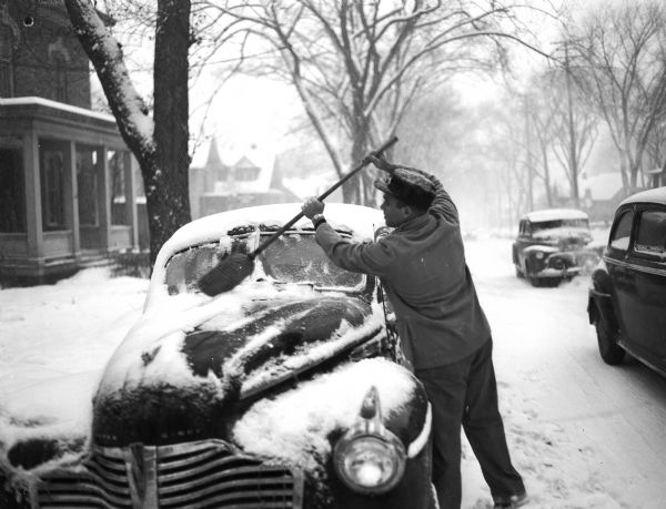 A winter scene of a man clearing his car windshield of snow at the corner of South Carroll Street and West Wilson Street, Madison, Wisconsin, after a winter storm that dropped eight inches of snow.