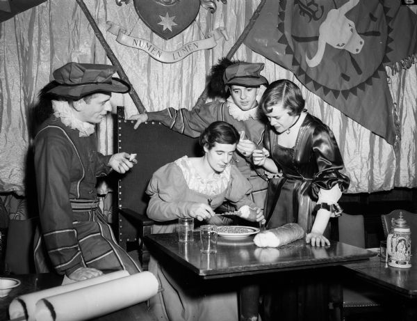 Nancy Berryman of Wauwatosa, (seated) is showing Clarence Bylsma, Racine; Henry Lippold, New London, and Anne Van Zandt, LaCrosse (left to right) all of whom are dressed in medieval costume, how to eat peas with bread and a knife, the only utensil used in the Tudor period, in preparation for the fifth annual Beefeaters' dinner held at the Memorial Union.