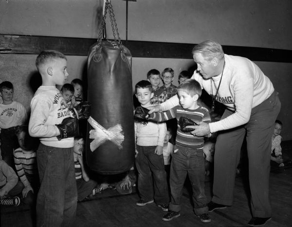 Instructor Verne Woodward (right), assistant boxing coach at the University of Wisconsin, instructs a group of small boys wearing boxing gloves on how to direct a punch at a punching bag. The classes were conducted at the Eagles Club Gym on Saturday mornings from December through Februrary since 1936. The boys, age six through fifteen, were divided into three age groups. From left, the boys are: Luke Lamboley, Jack Sweeney and John Yost. Other boys are watching from the background.