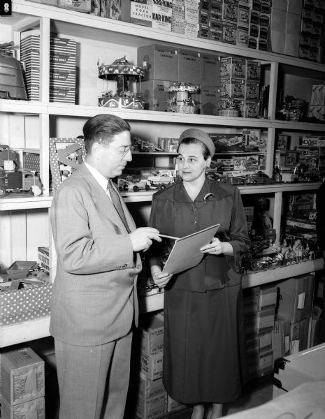 F.N. Weber and Mrs. William (Josephine) Doudna, head of the Empty Stocking Club toy distribution project, standing in front of shelves lined with toys.