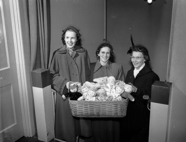 Three women are carrying a basket of dolls and teddy bears to be distributed to children through the Empty Stocking Club.  Left to right: Mrs. William (Shirley) Guelzow, Mrs. Walter (Marion) Thor, and Mrs. Lloyd (Celestine) Hein.