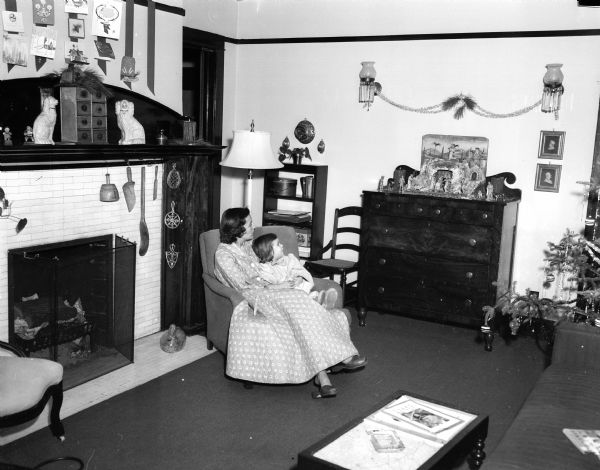 A mother and daughter from the Thomas family sit in a chair beside a fireplace while looking at a nativity scene arranged on a chest of drawers.