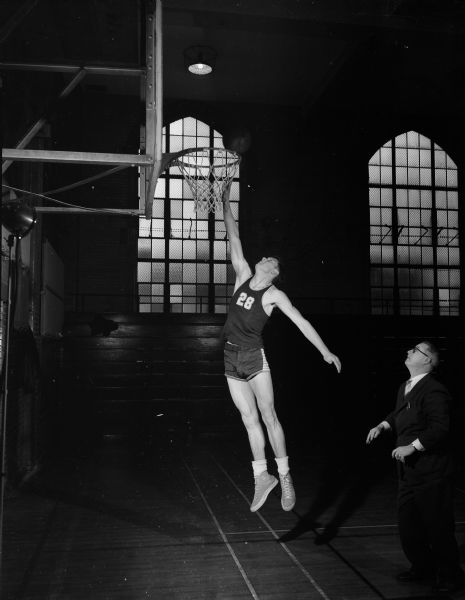 Charles Brendler, forward for the East High School basketball team, makes a shot at the basket. A man wearing a suit is standing on the right,