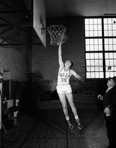 Owen Roberts, a forward on the Madison West High School basketball team and one of the team's three leading scorers, poses on the court. A man and a young boy are standing on the right watching.