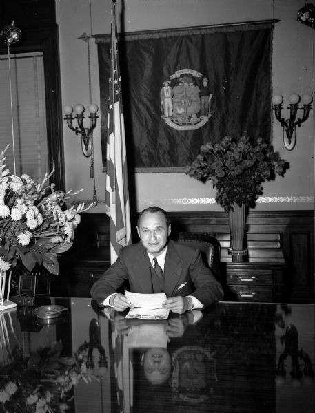 Newly inaugurated Governor Walter J. Kohler, Jr. sits at his desk in the Governor's office. Behind him are several bouquets of flowers and a Wisconsin flag.