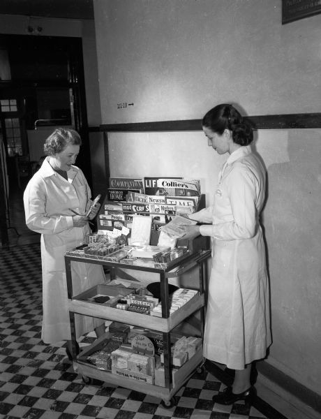 Madison General Hospital Auxiliary volunteer workers Edith Koehler, left, and Ellen Henderson checking the stock on the mobile cart containing magazines, cigarettes, small toilet articles, cosmetics, candy, and hand-knit baby gifts for sale to the patients.
