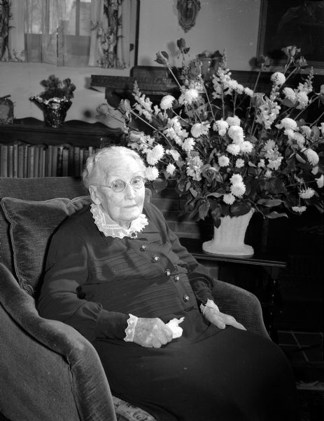 Portrait of Mrs. Jane McIlquaham sitting in an armchair on her 100th birthday. A floral arrangement is on a table behind her.