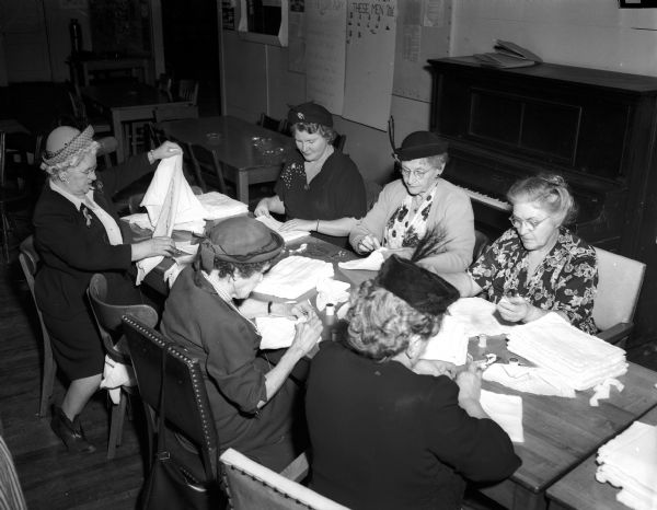 A group of six women sit around a table while sewing.