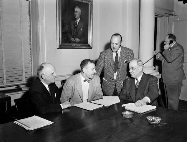UW football Coach Ivan "Red" Williamson accepts a new salary agreement on behalf of his assistants and himself. Pictured left to right, are: President E.B. Fred; Coach Williamson; Professor Nate Feinsinger, chairman of the Wisconsin athletic board, and A. Matt Werner, vice president of the Wisconsin board of regents. An unidentified man is standing while using a telephone in the background.