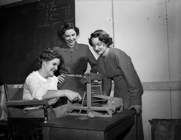 Two Junior Division University League members are shown with a trainee at the League's homecrafters' program  at Truax Field. Seated at a weaving loom is trainee Miss Patsy Collins. Standing left to right are  Mrs. Erich Farber and Mrs. Don (Jean) Voegeli.

