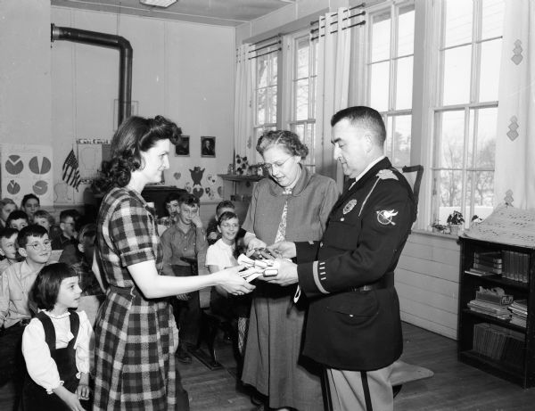 As Paoli grade school children watch, Lieutenant Arden C. Pope, head of the Dane County traffic safety division, presents badges and belts for school patrol boys and girls to teachers Mrs. Arthur Fahey (left) and Mrs. Una Hale.