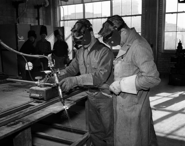 Two men wear welding masks while operating an acetylene cutting machine while three other students look on. Welding Instructor Fred Theiler (right) watches as student Dick Connery (left) learns to weld. They are in the newly completed vocational addition and participating in the very first year of Central High School, Trade and Technical Courses.