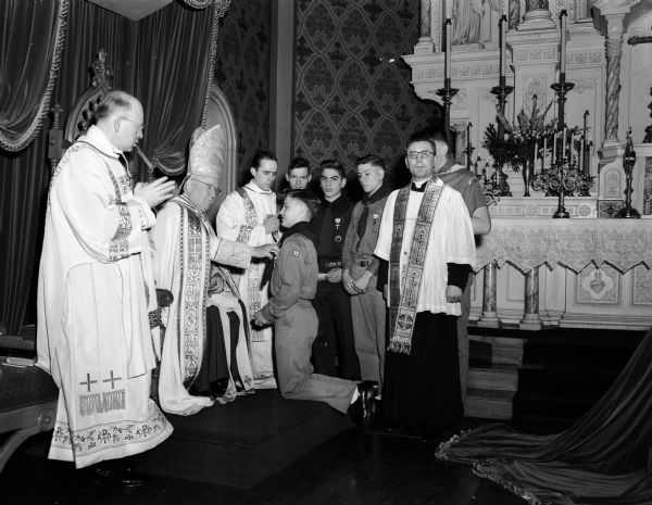 Five Catholic boy scouts from two Janesville parishes receiving the Ad Altare Dei Medal from Bishop William P. O'Connor of the Madison diocese in St. Raphael's Cathedral, Madison. Vincent McQuade kneels as the Bishop pins on his medal. Other scouts who received the award were Jerry Ford, Barry Kreft, Frank Rabiola, and Robert Stadler. Church dignitaries are (left to right): the Reverend Nicholas Schneider, deacon; Bishop O'Connor; the Reverend Wayne Turner; and the Reverend Josef V. Cieciorka, diocesan scout chaplain.