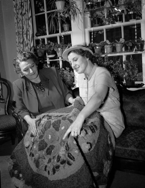 Portrait of two Catholic Women's Club members with their hobbies to be displayed at the Club's first hobby show. On the left is Mrs. Kathleen Huels who raises violets, some visible in the windows in the background. At the right is Mrs. Sophie Nichols with one of her hand-made rugs.