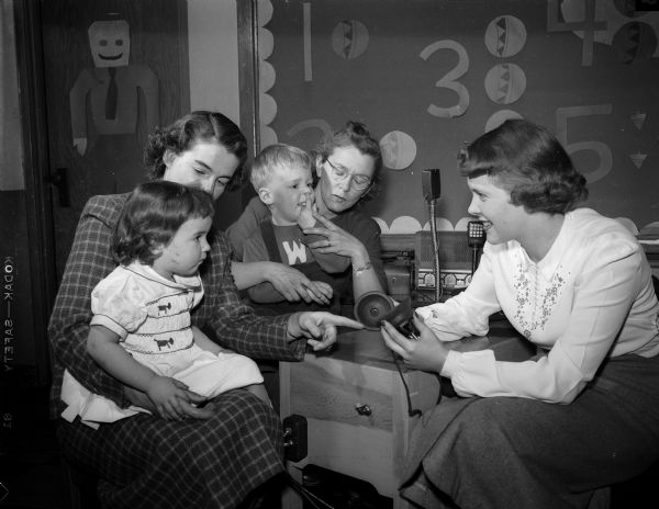 Two children with a hearing impairment, each seated in their mother's lap, learn about an instrument designed to help them hear. It will be used in an upcoming experimental workshop for hearing-impaired children under four years old. Left to right: Mrs. Allen (Alice) Stokes holds her daughter Susan, Mrs. Leo (Frances) Eberhardy and her child, and Lapham School kindergarten teacher, Ella Ziller.