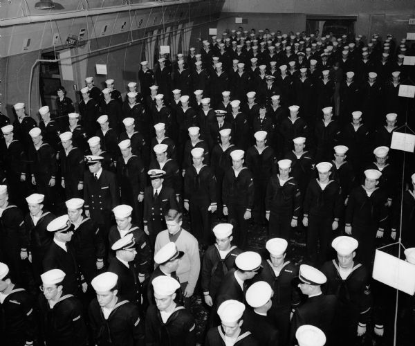 Elevated view of Madison's Organized Surface Division 9-212, naval reserves, being inspected at the naval reserve training center, located at 1046 East Washington Avenue. Heading the group of inspecting officers is (right to left): Lieut. Comdr. W.R. Kneisler, instructor-inspector at the Milwaukee training center; Comdr. W.M. Greeley, instructor at the Madison center; Lieut. C.H. Haynie, commanding Division 9-212; Lieut. E.A. Ruebel, inspector-instructor at the Kenosha center; Lieut. H.J. LeTart, assistant inspector-instructor at Milwaukee; and Lieut. Comdr. J.E. McCue, inspector-instructor at Madison.
