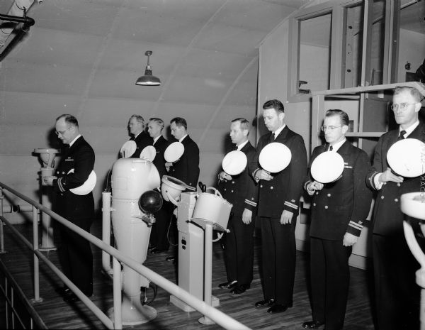 Group portrait of Wisconsin Naval Reserve Training Officers at the Madison naval reserve training center, located at 1046 East Washington Avenue, during an inspection of Madison's Organized Surface Division 9-212, naval reserves. Included are: Lieut. Comdr. W.R. Kneisler, instructor-inspector at the Milwaukee training center; Comdr. W. Greeley, instructor at the Madison center; Lieut. C.H. Haynie, commanding Division 9-212; Lieut. E.A. Ruebel, inspector-instructor at the Kenosha center; Lieut. H.J. LeTart, assistant inspector-instructor at Milwaukee; and Lieut. Comdr. J.E. McCue, inspector-instructor at Madison.