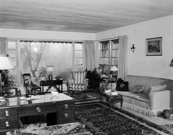Interior view of the living room at the John and Lulu Malin house, located at 3212 Topping Road in Shorewood Hills. The room is furnished with a sofa, upholstered chairs, and a desk; it is decorated with potted plants, a framed print or photograph, and multiple area rugs. A picture window looks out onto a residential area.