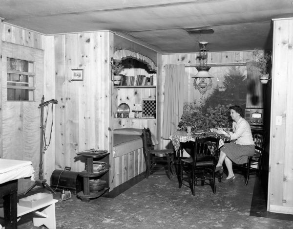 Lulu Malin sitting at a folding table in front of a picture window in the guest alcove at one end of the large recreation room in the John and Lulu Malin house in Shorewood Hills. On either side of the alcove are built-in bunk beds.