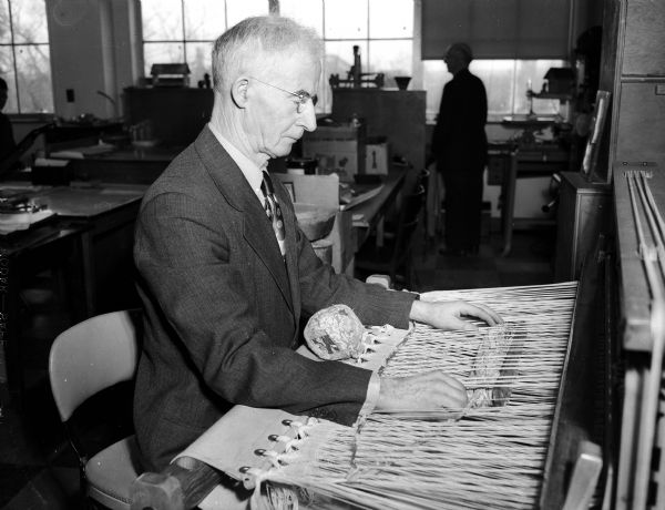 An unidentified man sits at a loom making a rug while receiving instruction in weaving as a participant in the Madison Vocational School home craft program. This program teaches home crafts to the disabled, tailoring the crafts to suit individual needs, with the goal of developing skills in the disabled so they can become self-supporting.