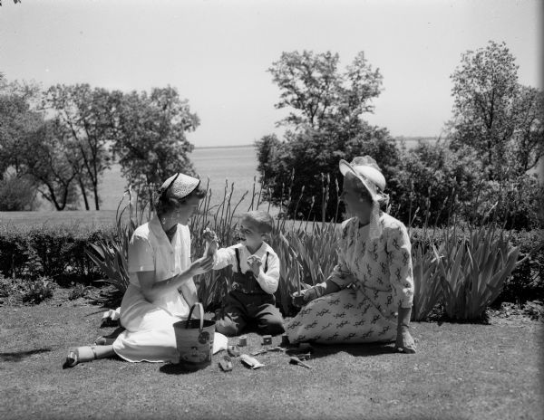 Mrs. Harley Graf and her hard-of-hearing son, Bobby, have lunch with Mrs. George C. (Dorothy) Guild on the lawn of Mrs. S.L. Odegard's residence on Lake Mendota at Fox Bluff. The occasion is the annual spring luncheon of the Attic Angel Association, one of the oldest philanthropic organizations in Wisconsin.
