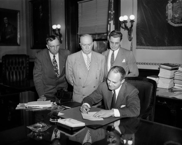 Governor Walter Kohler signs into law the bill providing that the governor and the legislature shall review all public expenditures, including money from revolving and segregated funds. Standing beside the governor as he signs the bill are, from left, Assemblyman Alfred R. Ludvigsen (R-Hartland), chairman of the assembly's finance committee;  Senator Foster B. Porter (R-Bloomington), senate finance commitee chairman; and Senator Arthur Lenroot (R-Superior), chairman of the budget committee of the legislative council.