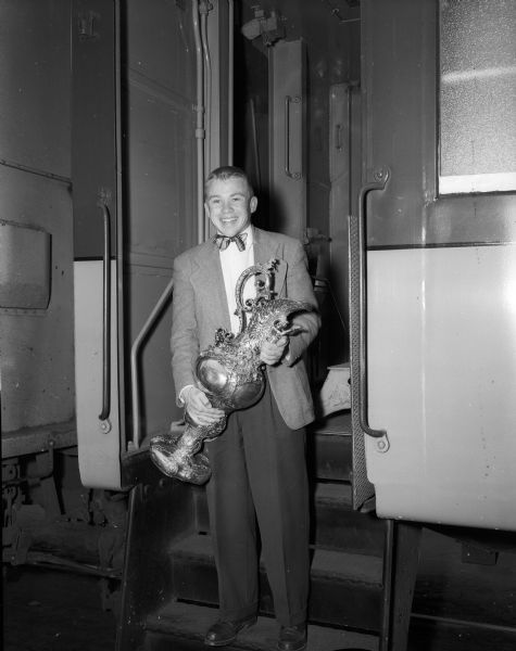 Captain Duane Daentl is holding the Varsity Challenge Cup that the rowing crew won at the Intercollegiate Rowing Association Championship races at Marietta, Ohio. He is shown wearing a bow tie and suit while standing on a step of a train car at the West Washington Avenue train station. The original caption reads: "A great little coxswain with a big trophy."
