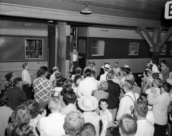 A crowd greets the University of Wisconsin varsity crew as they arrive on a train at the West Washington Avenue railroad station. At top center is crew captain and coxswain, Duane Daentl, holding the rowing trophy they won in Marietta, Ohio.