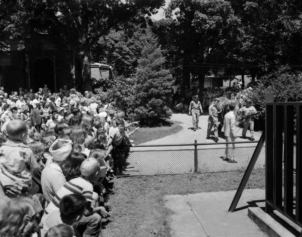 Winkie, Vilas Zoo elephant, is being led to her summer quarters at the zoo with a large crowd looking on. She is being led by attendants, Gordon Sheets and Dale Coyler, with Vilas Zoo (Vilas Park Zoo) director, Harold Hayes, looking on.