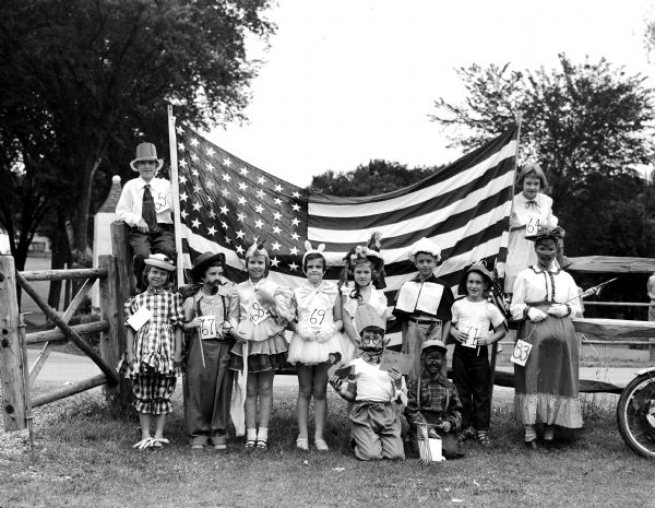 Twelve children from the Maple Bluff Fourth of July parade wearing varied costumes and standing in front of a large United States flag.