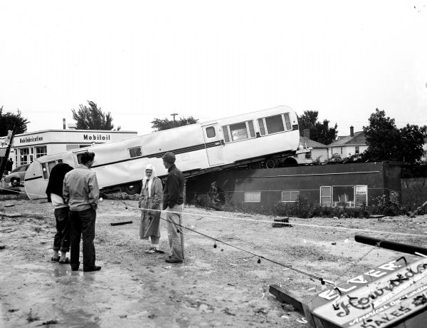 Four people are in the foreground overlooking two storm damaged large trailer homes, one piled on top of another. They and the trailers are at Elver Trailer Mart at 2945 E. Washington Avenue.  In the upper left corner is Arthur H. Stieve's Mobil Gas Station at 2951 E. Washington Avenue.