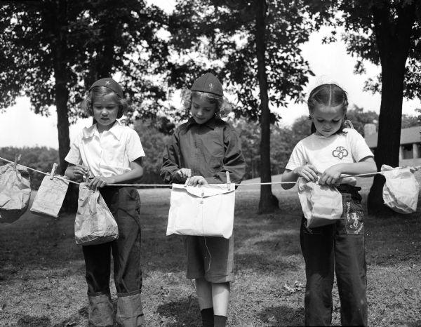 A scene at Brownie Day Camp at Hoyt or Burrows Park. Three Brownie Girl Scouts are hanging their "nosebag" lunches on a line. Left to right are: Mary Groth, Nancy Jo Terrance and Ellen Hafstad.