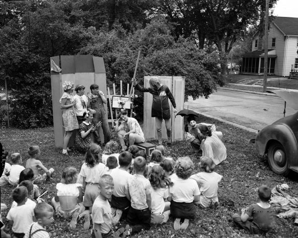 A group of children sitting on the lawn in a residental neighborhood watching seven other children acting in a play in front of a make-shift stage.