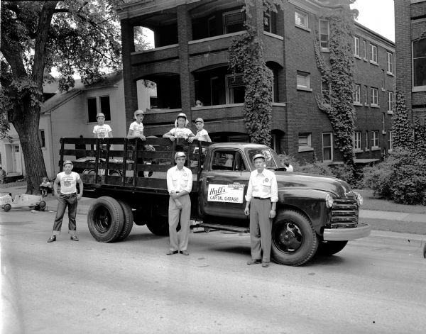 A pick-up truck from Hult's Capital Garage parks in front of 115 East Gorham Street with several derby car racers in the truck bed.