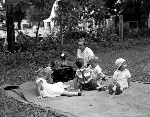 Six children are sitting on a canvas with Mrs. Ralph Huitt while listening to a record at U.W. Houses Outdoor Nursery and Play School. Children include Cynthia Huitt, Johnny Wilde, Jimmy Gilchrist, Maureen White, Patty McDonald, and Carol Duncan.