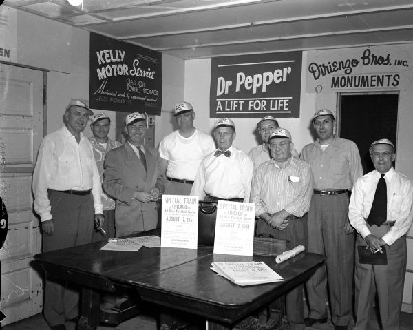 A group of nine men, members of the Westside Business Men's Association, standing around a table with advertisements for a "Special Train to Chicago" for an all star football game. The men are wearing souvenir caps. There are signs for Kelley Motor Services, Doctor Pepper, and Direnzo Bros., Inc. Monuments.