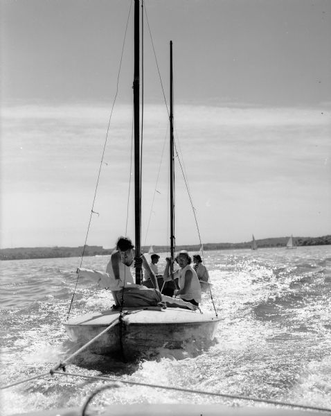 Four youth, members of the Mendota Yacht Club, in a sailboat on Lake Mendota. The sailboat is being towed by another boat. Other sailboats can be seen in the distance.
