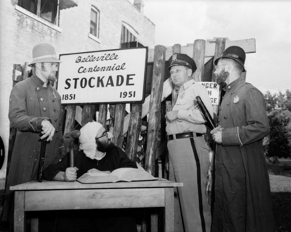 Four men pose in front of the "Belleville Centennial Stockade, 1851-1951." Two are dressed as old time police officers and one is dressed as a judge. They are sentencing a man dressed in a contemporary police officer's uniform. The celebration was held September 1-3, 1951.