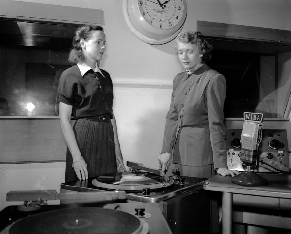 Frances Hurst (left) and Marcella Gill (right) listen to voice recordings at the WIBA radio broadcasting studio. Hurst was chairman of the WHA radio programs for the league. Gill was president and moderator for a second League radio program at WIBA on local issues. Two large phonograph record players stand in the foreground. Also visible are a WIBA microphone and other radio broadcasting equipment.
