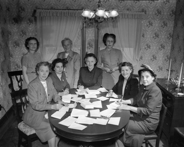 Officers of the Catholic Women's Club gather around a table while preparing yearbooks for mailing. Standing, from left to right, are: Mrs. Wilbur R. Carnes, treasurer; Mrs. Roy L. Hilsenhoff, president; and Mrs. Leo Dwyer, auditor. Seated, left to right are: Mrs. Ben F. Rusy, second vice-president; Mrs. Joseph Gruno, first vice-president; Mrs. Earl Wilke, historian; Mrs. Lyle Minto, corresponding secretary; and Mrs. John L. Nichols, recording secretary.